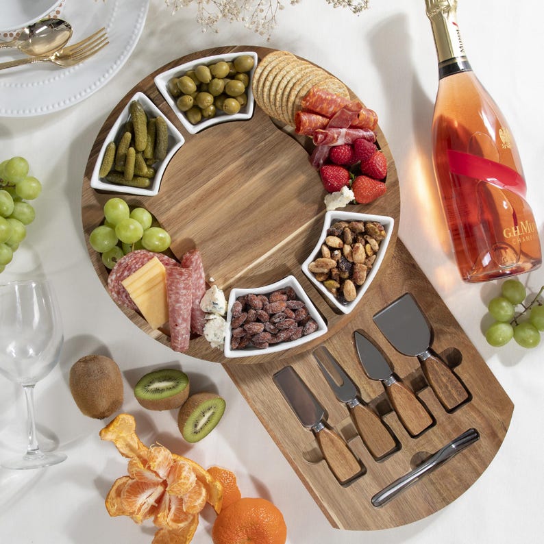 May include: A wooden charcuterie board with a circular design, featuring small white bowls filled with olives, pickles, and nuts. The board is laden with crackers, meats, and fruit, alongside a bottle of ros&eacute; wine and cheese knives.