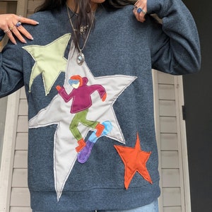 May include: A dark blue sweatshirt with a large white star featuring a cartoon figure snowboarding. The figure is wearing a purple jacket, green trousers, and a blue helmet. There are two smaller stars, one green and one orange, on the sweatshirt.