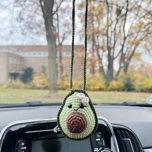Könnte beinhalten: Ein handgehäkelter Avocado-Autoanhänger. Die Avocado ist grün mit einem braunen Kern und einer kleinen rosa Blume. Der Anhänger hängt an einer grünen Kordel, die an einem Rückspiegel befestigt ist.