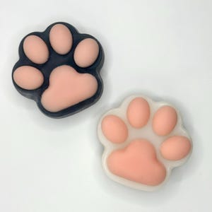 May include: Two paw-shaped soaps. One is dark gray with light pink pads, the other is white with light pink pads. The soaps are on a white background. The paw pads are rounded and smooth.