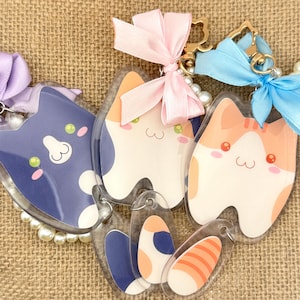 May include: Three clear acrylic keychains with cartoon animal designs. The keychains feature a blue cat, a white cat, and an orange cat. Each keychain has a pearl-like bead chain and a gold metal ring.