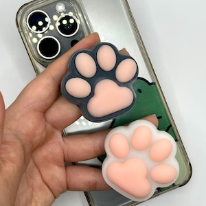 May include: Two paw-shaped phone grips. One is dark gray with pink pads, the other is white with pink pads. The grips are on a clear phone case with a green design.