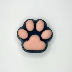 May include: A paw-shaped soap with a dark blue border and light pink pads. The soap is a close-up shot against a white background. The paw print design is a common theme for pet-related products.