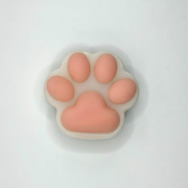 May include: A close-up of a paw-shaped soap with a light pink paw print design. The paw print is surrounded by a white border. The soap is on a white background.