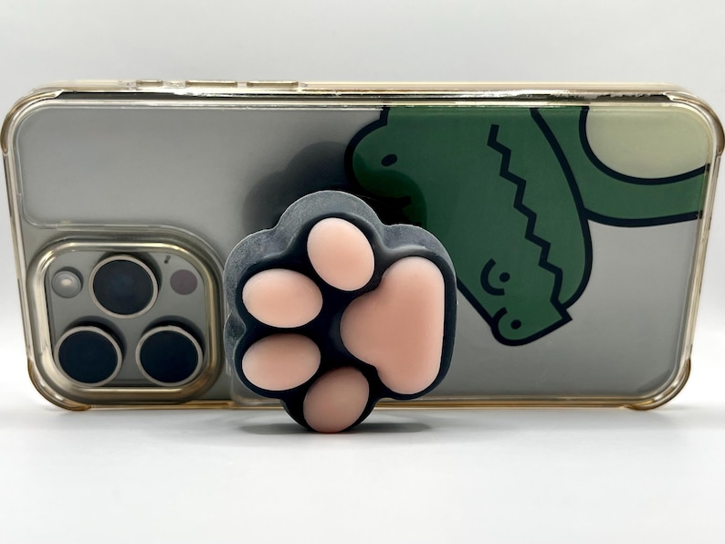 May include: A black and pink paw-shaped phone grip is attached to a clear phone case. The phone case has a green cartoon design on the back. The phone has a gold-colored frame and a triple-lens camera.