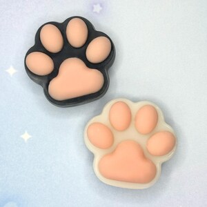 May include: Two paw-shaped decorative items. One is black with peach-colored pads, the other is white with peach-colored pads. The items are likely for crafts or embellishments, featuring a cute, animal-themed design.