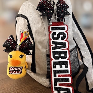 May include: A silver glitter backpack featuring a black and red bow keychain, a yellow duck plush keychain with the text COURT STARS, and a long vertical black and red glitter name tag with the name ISABELLA in white block letters.