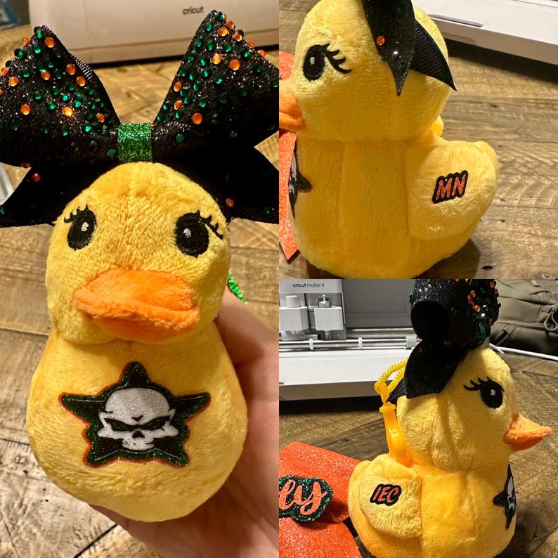 May include: Yellow plush duck with an orange beak and black eyes. The duck has a black bow with green and orange embellishments. A skull and crossbones design is on the duck's chest. The duck has the letters "MN" on its side.