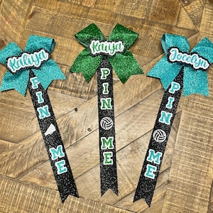 Puede incluir: Tres lazos de animadora brillantes con nombres personalizados. Un lazo es verde, dos son turquesas. Cada lazo tiene una cinta negra con las palabras "PIN ME" y un gráfico de voleibol. Los nombres "Kaliya" y "Jocelyn" son visibles.