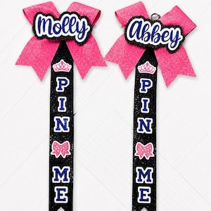 Può includere: Due fiocchi da cheerleader con i nomi "Molly" e "Abbey" in corsivo bianco su sfondo nero e blu. I fiocchi sono glitterati rosa con dettagli neri e blu. I nastri riportano la scritta "PIN ME" con una corona e un fiocco.