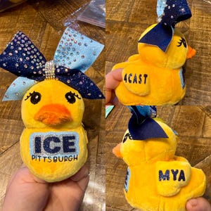 May include: Yellow plush duck with a large blue and silver glitter bow. The duck has an orange beak and black eyes. The text "ICE PITTSBURGH" is on the front. Another duck has the text "4CAST" and a third has "MYA".