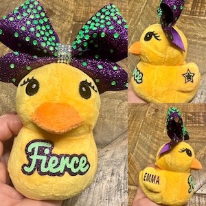 May include: Yellow plush duck with an orange beak and eyelashes. The duck has a purple and green glitter bow. One duck has the word "Fierce" in green. Another has the word "Emma". A third has the word "Ice" and a star.