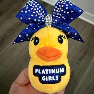 May include: A yellow plush duck toy with an orange beak and black eyes. The duck has a large blue glitter bow adorned with rhinestones. The text "PLATINUM GIRLS" is printed on the duck's chest.