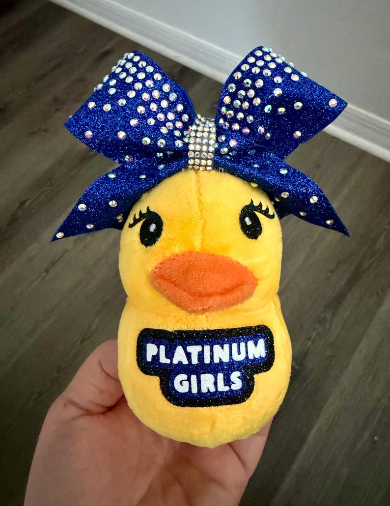May include: A yellow plush duck toy with an orange beak and black eyes. The duck has a large blue glitter bow adorned with rhinestones. The text "PLATINUM GIRLS" is printed on the duck's chest.