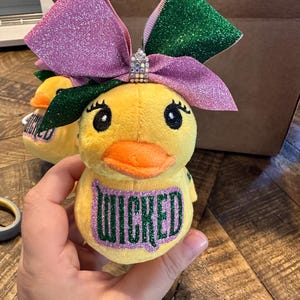May include: Yellow plush duck toy with an orange beak and large eyes. The duck has a large glittery bow in shades of pink and green. The word "WICKED" is printed on the duck's chest in green glitter.