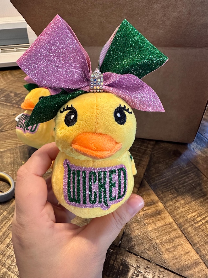 May include: Yellow plush duck toy with an orange beak and large eyes. The duck has a large glittery bow in shades of pink and green. The word "WICKED" is printed on the duck's chest in green glitter.