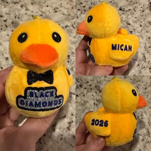 May include: A plush yellow duck with an orange beak and black eyes. The duck has a black glitter bow tie and the words "BLACK DIAMONDS" in a blue and silver design. The name "MICAH" is embroidered on the side, and the number "2026" is on the back.