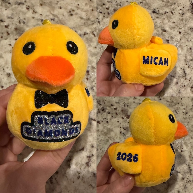 May include: A plush yellow duck with an orange beak and black eyes. The duck has a black glitter bow tie and the words "BLACK DIAMONDS" in a blue and silver design. The name "MICAH" is embroidered on the side, and the number "2026" is on the back.