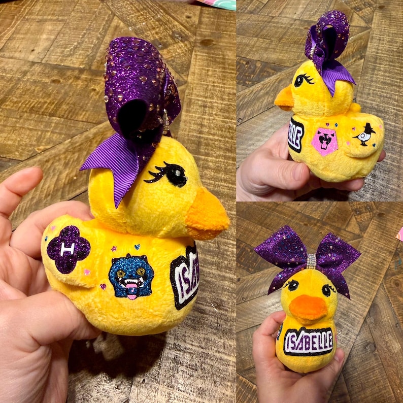 May include: Yellow plush duck with a purple glitter bow and the name "Isabelle". The duck has an orange beak and black eyes. It features embroidered patches with a letter "H" and a monster design.