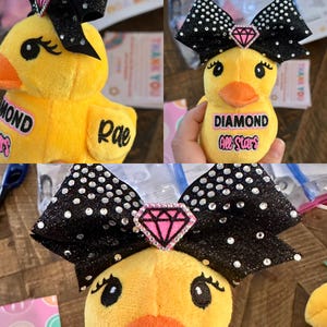 May include: A plush yellow duck with an orange beak and black eyes. The duck has a large black bow with silver glitter and a pink diamond-shaped embellishment. The text "DIAMOND All Stars" is on the duck's body.