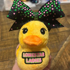 May include: A yellow plush duck with an orange beak and black eyes, wearing a black bow with green and pink polka dots. The duck has the text "LONESTAR LADIES" on its chest.