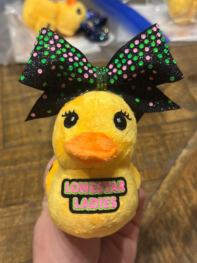 May include: A yellow plush duck with an orange beak and black eyes, wearing a black bow with green and pink polka dots. The duck has the text "LONESTAR LADIES" on its chest.
