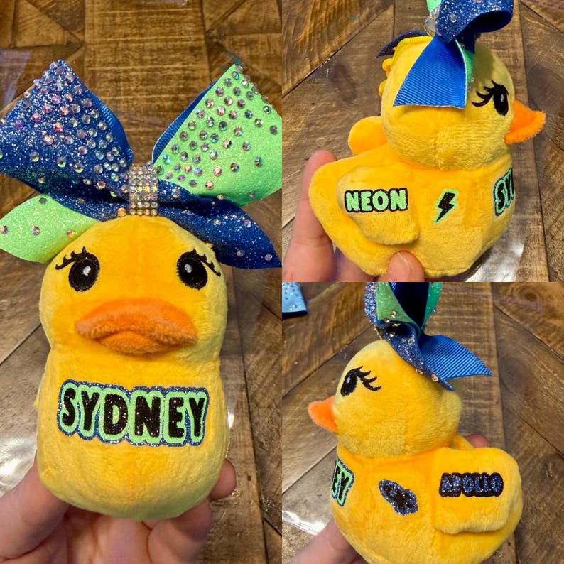 May include: Yellow plush duck with a blue and green glitter bow. The duck has an orange beak and black eyes. The name "SYDNEY" is embroidered on the front. Other ducks have the names "NEON" and "APOLLO".