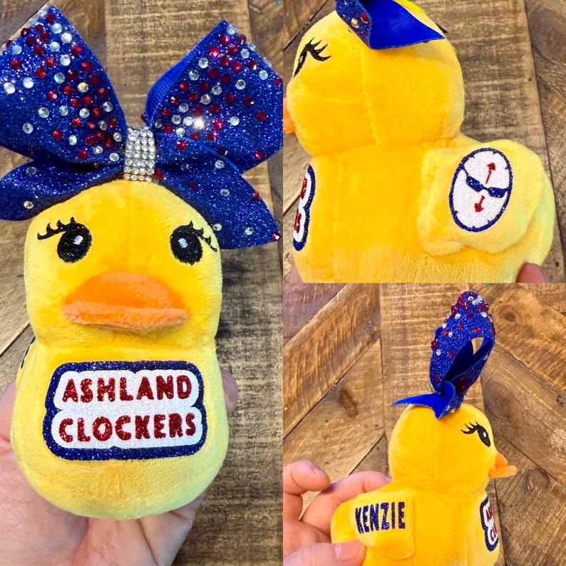 May include: A plush yellow duck with a blue and red glitter bow. The duck has an orange beak, black eyes, and the text "ASHLAND CLOCKERS" on its belly. The duck also has the name "KENZIE" embroidered on a yellow tag.