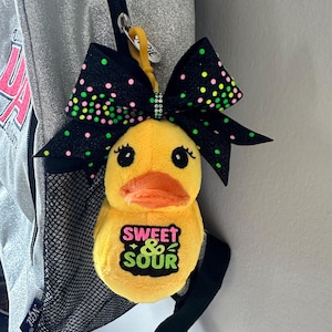 May include: A yellow plush duck keyring with an orange beak and black eyes. The duck has a green and black bow with pink and green polka dots. The text "SWEET & SOUR" is on the duck's chest.
