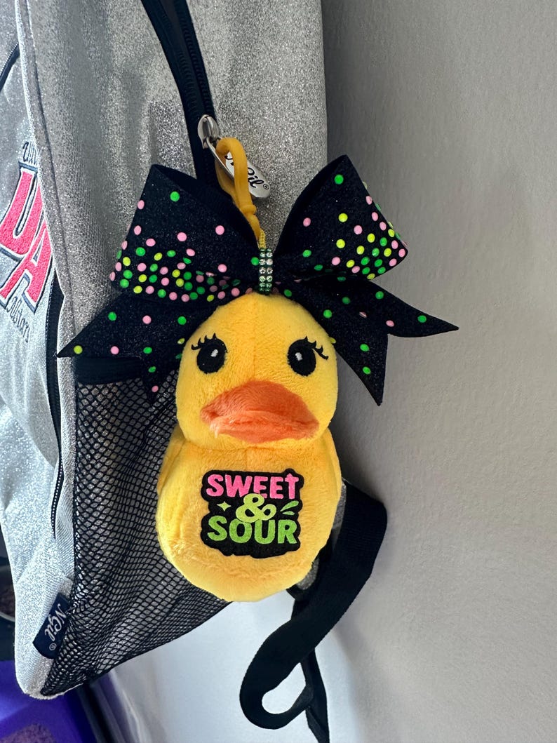 May include: A yellow plush duck keychain with an orange beak and black eyes. The duck has a green and black bow with pink and green polka dots. The text "SWEET & SOUR" is on the duck's chest.