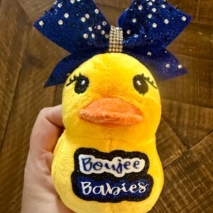 May include: A yellow plush duck toy with a blue glitter bow and a rhinestone accent. The duck has a bright orange beak and the text "Boujee Babies" on its belly.