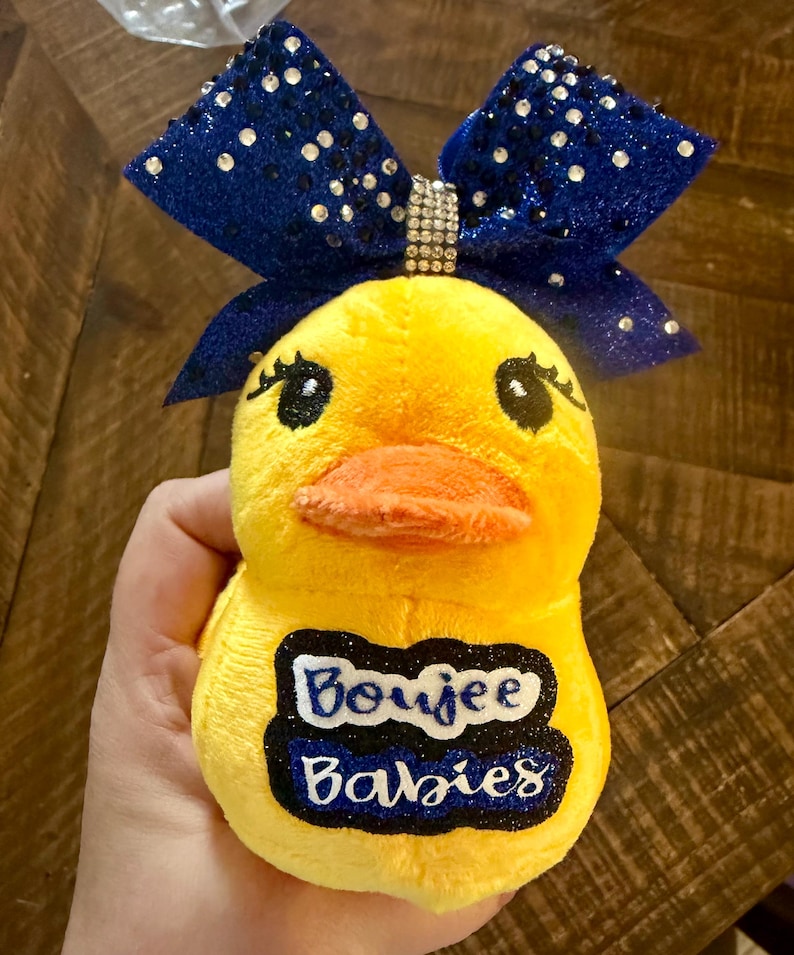 May include: A yellow plush duck toy with a blue glitter bow and a rhinestone accent. The duck has a bright orange beak and the text "Boujee Babies" on its belly.