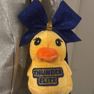 May include: A yellow plush duck with an orange beak and a blue glitter bow. The duck has the text "THUNDER ELITE" in blue and gold on its chest. The duck is attached to a yellow clip that is attached to a backpack.