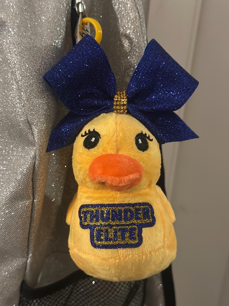 May include: A yellow plush duck with an orange beak and a blue glitter bow. The duck has the text "THUNDER ELITE" in blue and gold on its chest. The duck is attached to a yellow clip that is attached to a backpack.