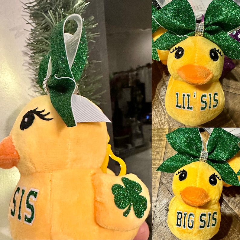 May include: Yellow plush duck ornament with a green glitter bow and a white ribbon. The duck has black eyelashes and the word "SIS" on its side. A green glitter shamrock is on the duck's side. The text "LIL' SIS" and "BIG SIS" are on the duck's belly.