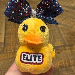 May include: A yellow plush duck toy with a navy blue glitter bow adorned with red and crystal rhinestones. The duck has an orange beak, black eyes, and the word "ELITE" is on the chest in navy with a white and red offset. A fun backpack accessory.