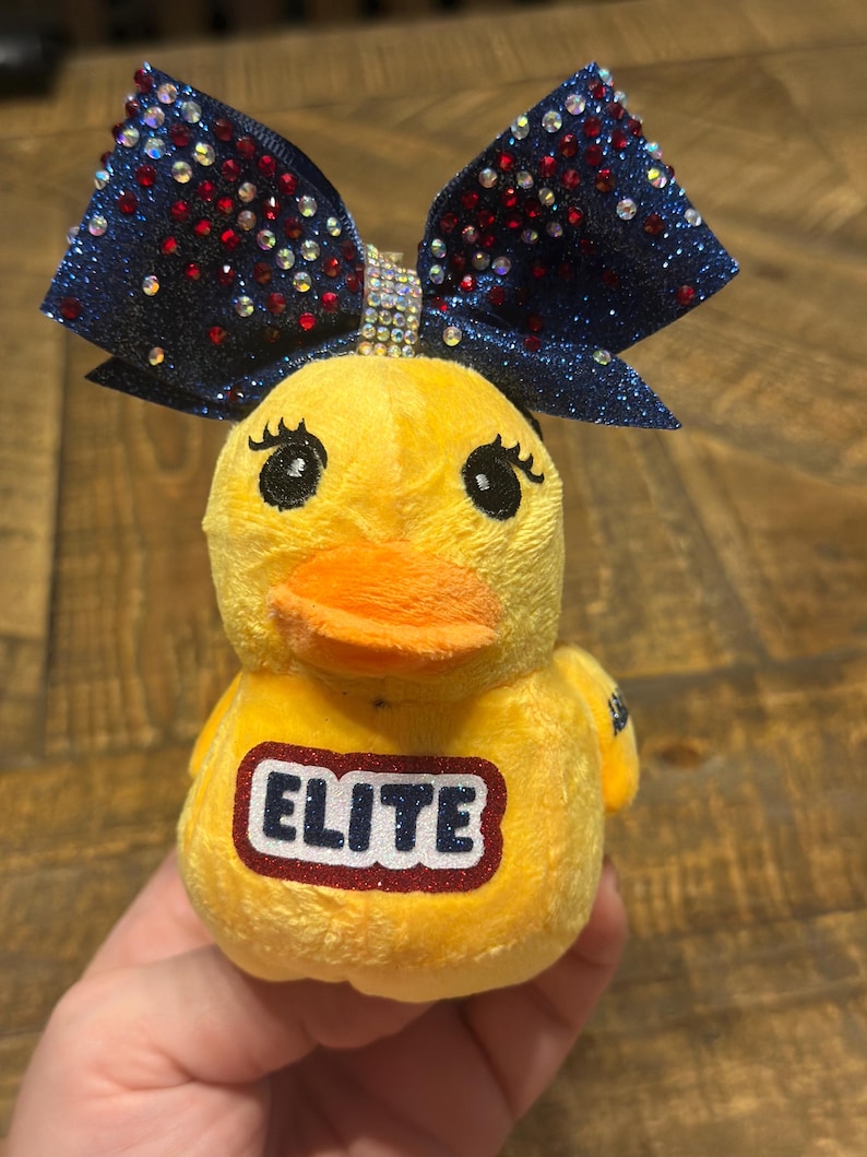 May include: A yellow plush duck toy with a navy blue glitter bow adorned with red and crystal rhinestones. The duck has an orange beak, black eyes, and the word "ELITE" is on the chest in navy with a white and red offset. A fun backpack accessory.