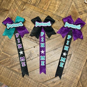 May include: Three glittery cheerleading bows with the name "Savannah" on top. Each bow has a different color: teal, black, and purple. Below the bow, the words "PIN ME" are spelled out vertically with a star.