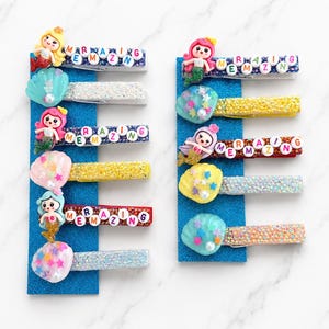 May include: A collection of mermaid-themed hair clips. Each clip features a mermaid figure, the word "AMAZING", and decorative elements like seashells and glitter. The clips are arranged on blue glittery backing.