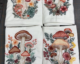 Op de afbeelding: Vier witte keukendoeken met geborduurde paddenstoelontwerpen. Elke doek heeft een andere opstelling van kleurrijke paddenstoelen, bloemen en bladeren in tinten rood, oranje, bruin en groen.