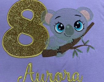 Könnte beinhalten: Ein lila T-Shirt mit einer goldenen Glitzerzahl 8, einem gestickten Koalabären auf einem Ast und dem Namen "Aurora" in goldener Glitzer.