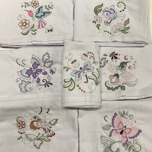 May include: Set of six white tea towels with embroidered floral and bird designs. The designs feature butterflies, hummingbirds, and floral motifs in shades of purple, pink, green, and brown.