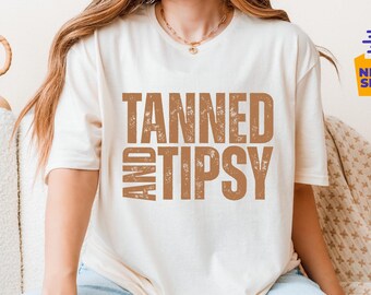May include: Cream-colored t-shirt with the words "TANNED AND TIPSY" in a distressed brown font. The shirt is a relaxed fit and is being worn. The image also includes a decorative vase and a pillow.