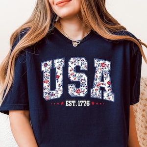 May include: A navy blue t-shirt with a floral print of the word "USA" and the text "EST. 1776" underneath.