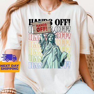 May include: A white t-shirt with a graphic of the Statue of Liberty holding a sign that says "Hands Off!" The graphic is surrounded by the words "Hands Off!" in a rainbow of colors.