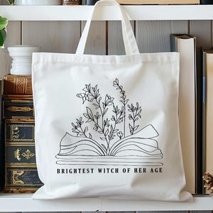 May include: White canvas tote bag with a black line drawing of an open book with flowers growing out of it. The text "Brightest Witch of Her Age" is printed below the book.