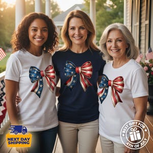 May include: Three short-sleeved t-shirts featuring a large graphic of a bow with a red, white, and blue American flag pattern. Two shirts are white and one is navy blue. The bow design includes white stars on the blue section and red stripes.