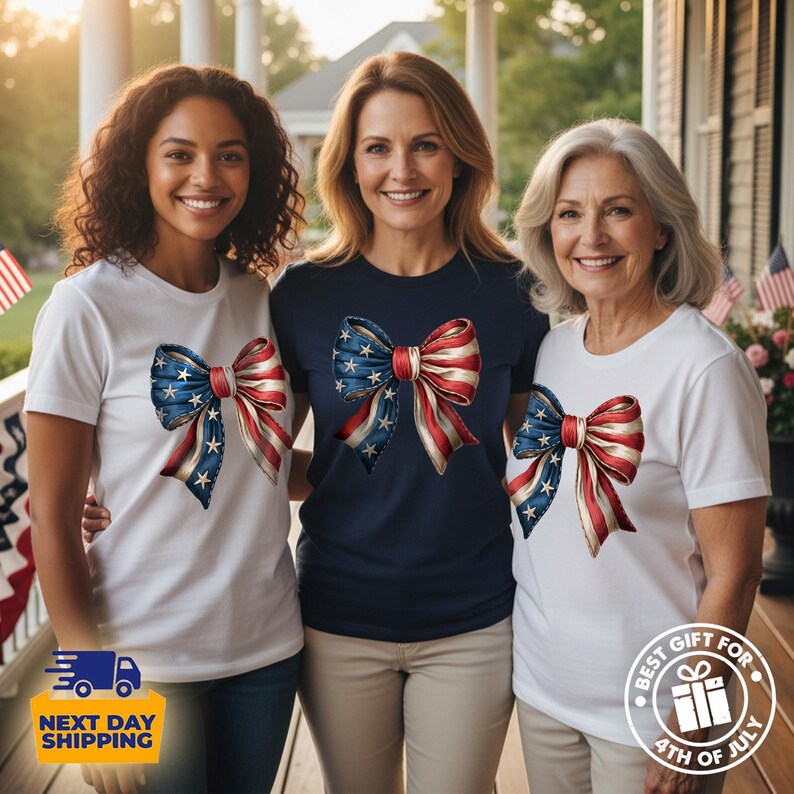 May include: Three short-sleeved t-shirts featuring a large graphic of a bow with a red, white, and blue American flag pattern. Two shirts are white and one is navy blue. The bow design includes white stars on the blue section and red stripes.