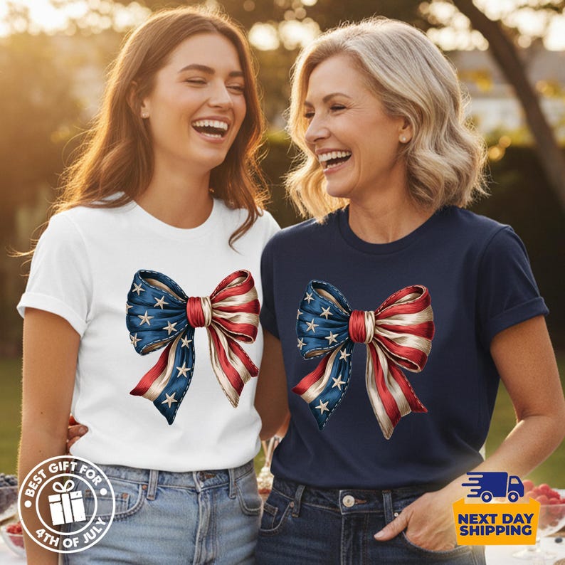 May include: A white t-shirt and a navy blue t-shirt, each featuring a graphic print of a large bow decorated with the American flag pattern, including blue sections with white stars and red and white stripes on the fabric of the bow.