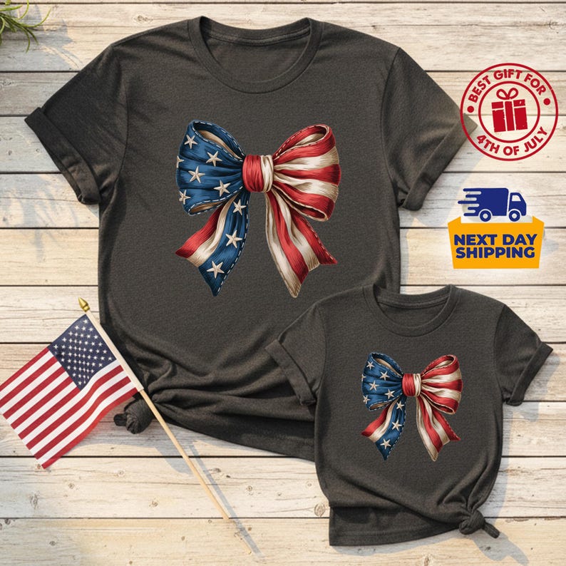 May include: Two charcoal gray short-sleeve t-shirts featuring a graphic of a large bow with a red, white, and blue American flag pattern. The bow has white stars on the blue side and red and white stripes on the other side. A small American flag is included.